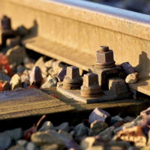 Close-up of railway tracks with rusty screws and fastening plates holding the rails to the wooden sleepers. There is ballast around the rails